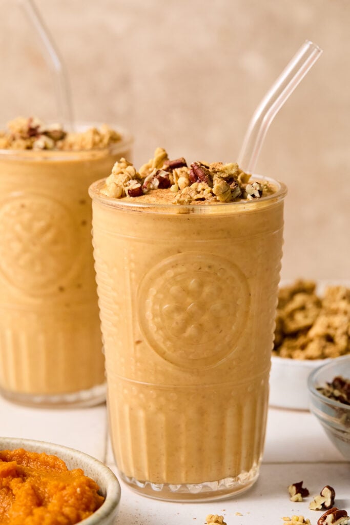 Pumpkin protein shake in a glass topped with chopped pecans and cinnamon, with a straw in it. Around the glass are some chopped pecans, a bowl of pumpkin puree, a bowl of chopped pecans, a bowl of granola, and another glass with a smoothie.