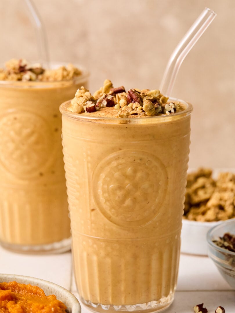 Pumpkin protein shake in a glass topped with chopped pecans and cinnamon, with a straw in it. Around the glass are some chopped pecans, a bowl of pumpkin puree, a bowl of chopped pecans, a bowl of granola, and another glass with a smoothie.