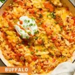 Buffalo chicken casserole in a large white enameled skillet topped with melted cheese, a dollop of greek yogurt, sliced green onions, and hot sauce. A kitchen towel is tied around the handle. Around the pan is a plate with two white forks, a small dish of sliced green onions, a bowl of shredded cheese, and another plate.