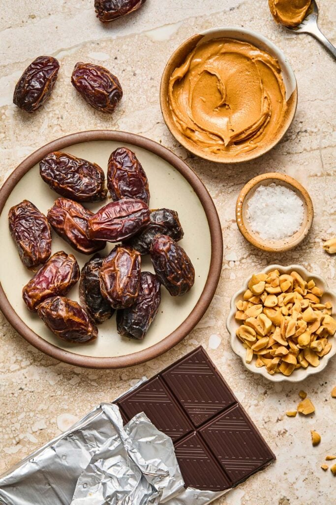 A spoonful of peanut butter, a bowl of creamy peanut butter, a small bowl of flaky sea salt, a bowl of chopped peanuts, a dark chocolate bar halfway unwrapped, a plate of medjool dates, and a few dates on a tan concrete counter.