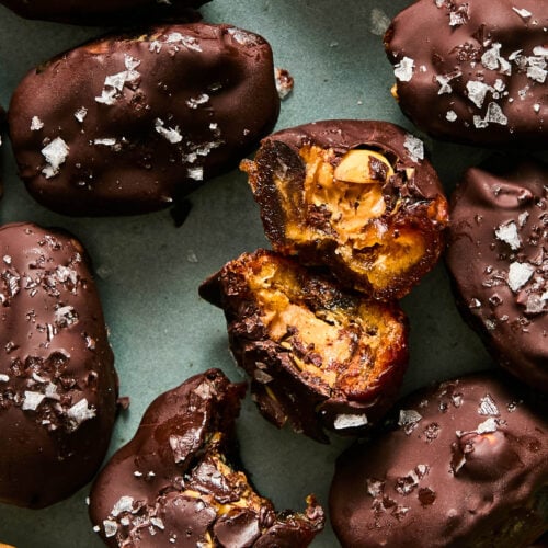 A plate of chocolate covered date snickers topped with flaky salt. One of the dates is broken up with the inside showing, and another has a bite taken out of it.