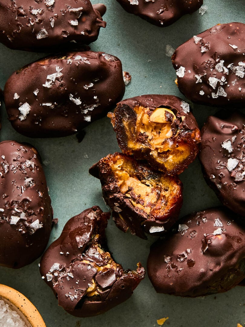 A plate of chocolate covered date snickers topped with flaky salt. One of the dates is broken up with the inside showing, and another has a bite taken out of it.