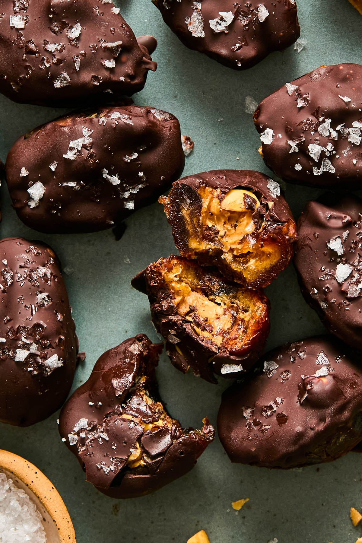 A plate of chocolate covered date snickers topped with flaky salt. One of the dates is broken up with the inside showing, and another has a bite taken out of it.