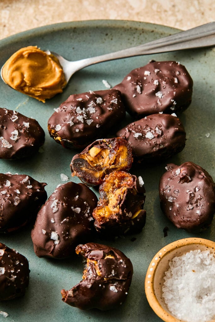 A plate of chocolate covered snickers dates topped with flaky salt. One of the dates is broken in half and standing up so you can see the inside, and another has a bite taken out of it. There is also a small dish of flaky salt and a spoonful of peanut butter on the plate.