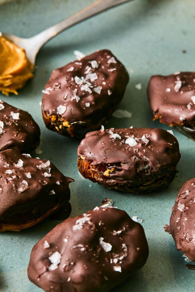 Chocolate covered snickers dates topped with flaky sea salt on a plate. A spoon with peanut butter is also on the plate.