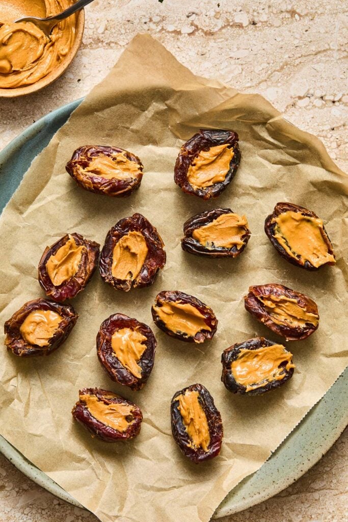 A parchment paper lined plate with a bunch of medjool dates that have been stuffed with peanut butter. Next to the plate is a bowl of peanut butter with a spoon in it.