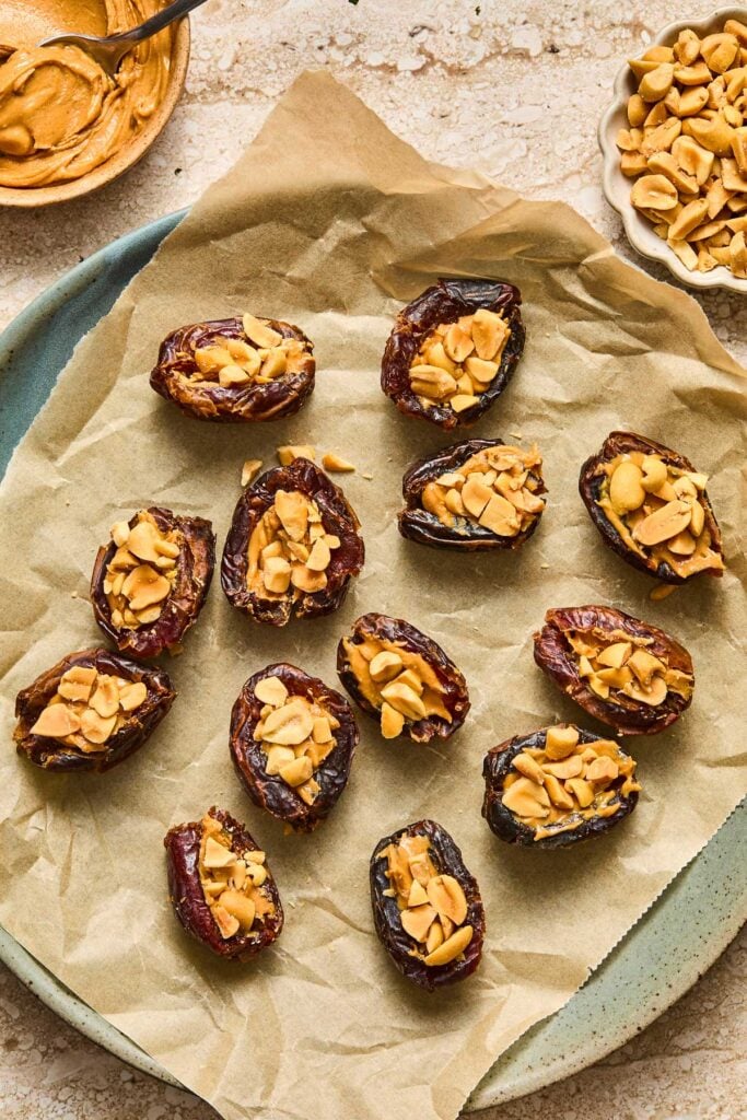 A parchment paper lined plate with a bunch of medjool dates that have been stuffed with peanut butter and chopped peanuts. Next to the plate is a bowl of peanut butter with a spoon in it and a bowl of peanuts.
