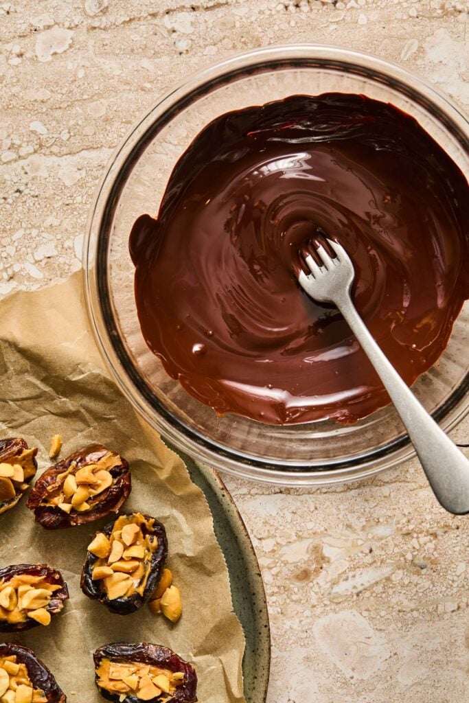 A bowl of melted chocolate with a fork in it. Next to the bowl is a parchment paper lined plate with dates stuffed with peanut butter and chopped peanuts.