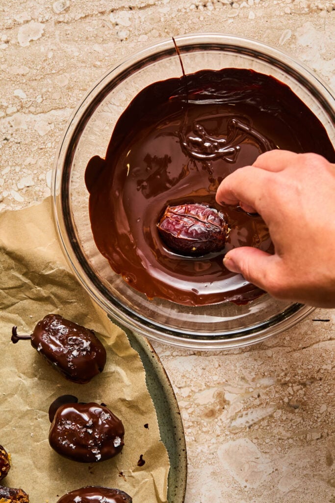 A bowl of melted chocolate with a stuffed date being dipped into it. Next to the bowl is a parchment paper lined plate with more chocolate covered stuffed dates.
