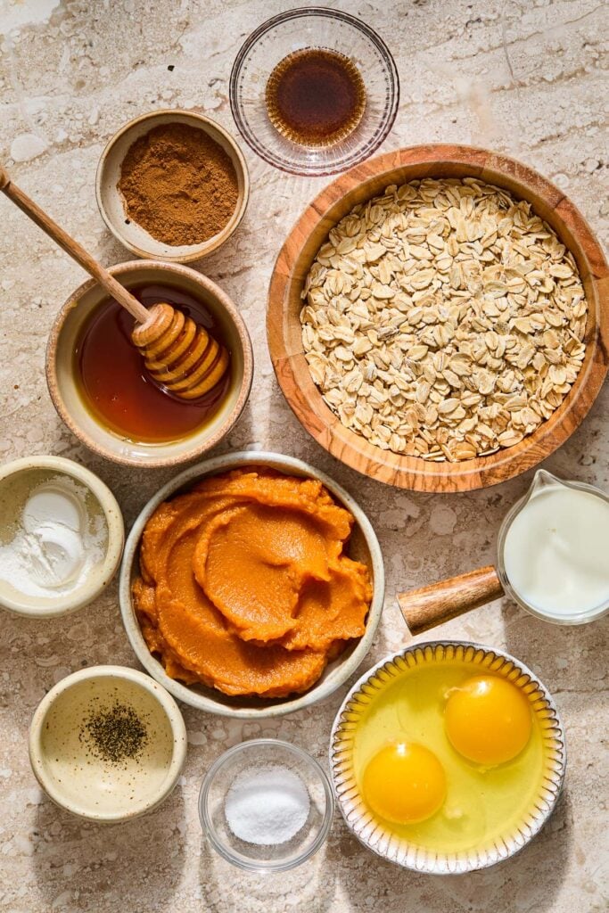 A small bowl of vanilla bean paste, a bowl of rolled oats, a measuring cup of milk, a bowl with two cracked eggs in it, a small bowl of salt, a bowl of pumpkin puree, a small bowl with a pinch of black pepper, a small bowl of baking powder, a bowl with honey and a honey dipper in it, and a bowl of pumpkin pie spice on a tan concrete counter.