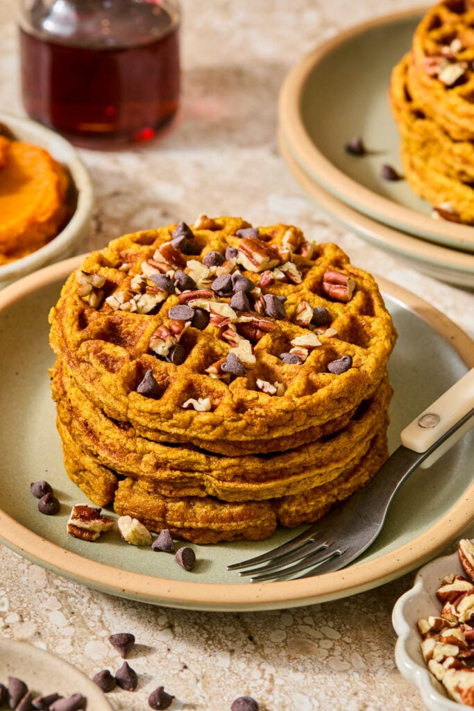 A plate of three pumpkin waffles stacked on top of each other topped with chocolate chips and chopped pecans. Around the plate is a bowl of chopped pecans, some scattered chocolate chips, a small bowl of pumpkin puree, a pitcher of maple syrup, and a plate with another stack of waffles on it.