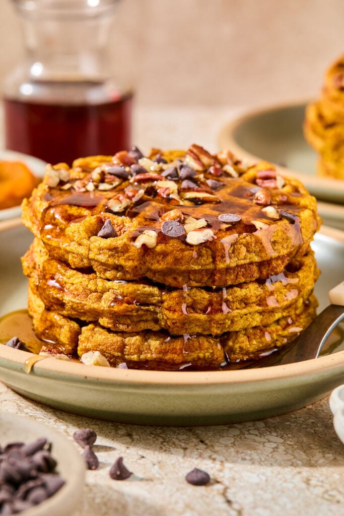 A stack of three pumpkin waffles topped with chocolate chips, chopped pecans, and maple syrup. Around the plate is a bowl of chocolate chips, a small bowl of pumpkin puree, a pitcher of maple syrup, and a plate with another stack of waffles on it.